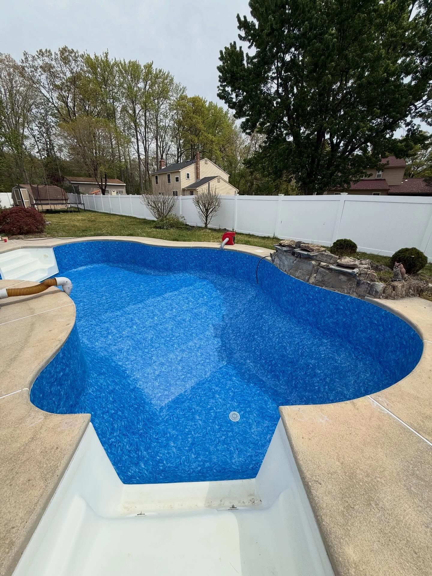 Backyard swimming pool with bright blue water, white fence, and surrounding patio and trees.