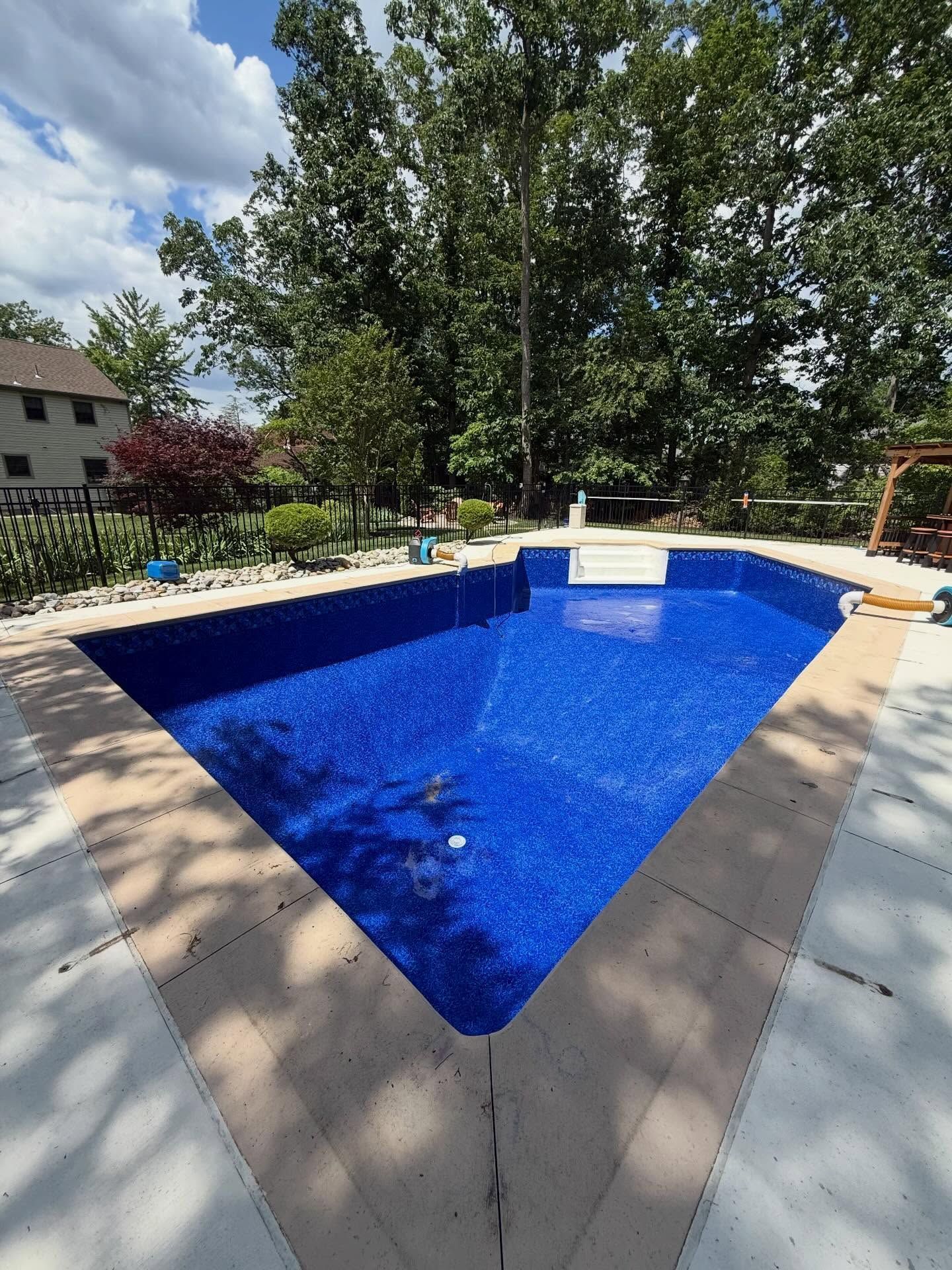 Backyard swimming pool with bright blue water, beige deck, and trees in the background