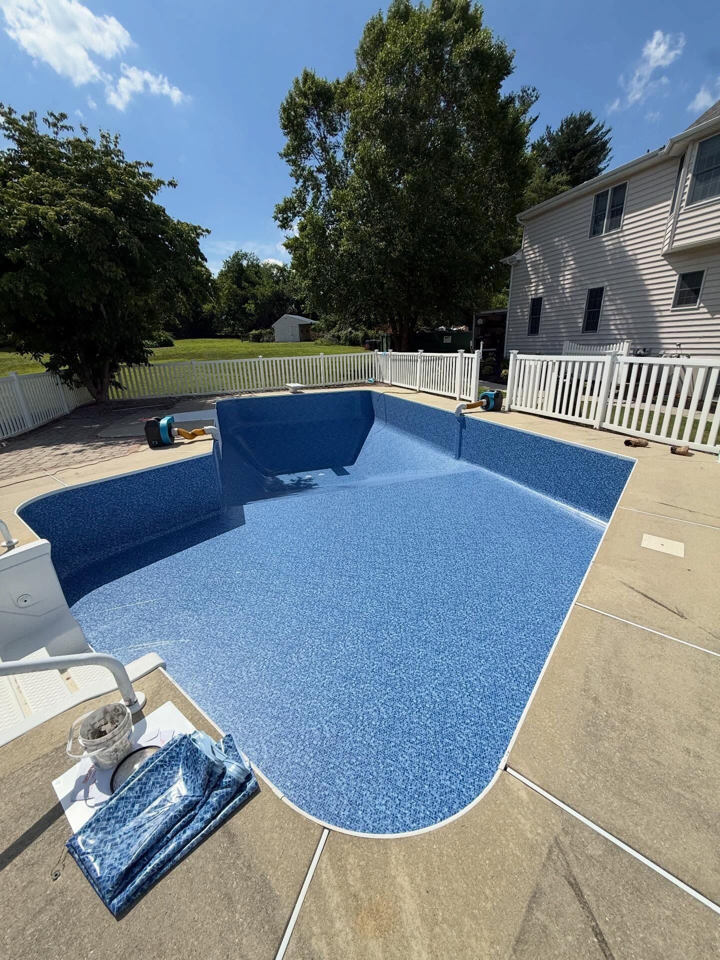 Backyard swimming pool with blue mosaic water, beige deck, white fence, and house on the right