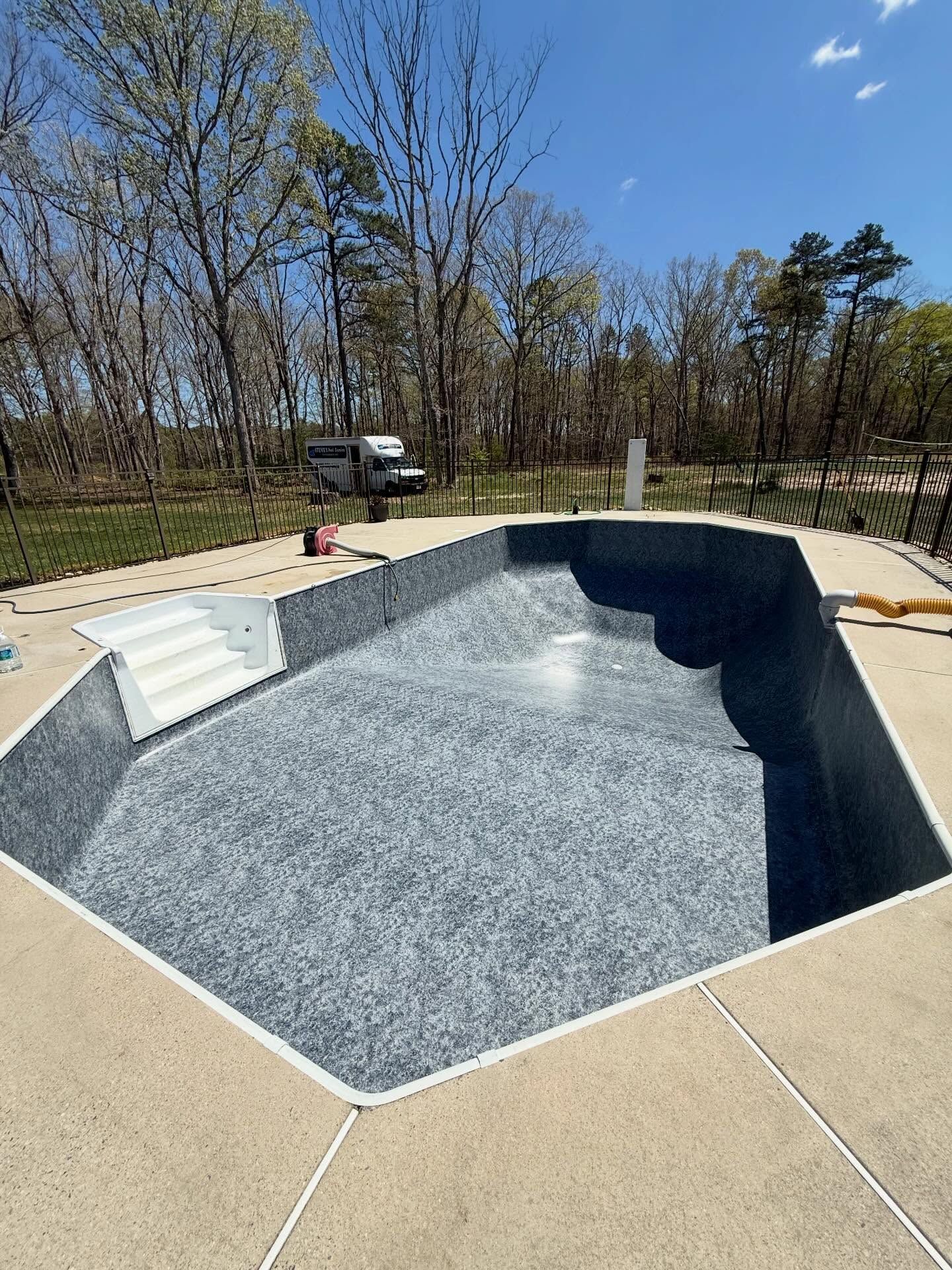 Empty in-ground swimming pool with blue interior, surrounded by a concrete deck and trees in the background