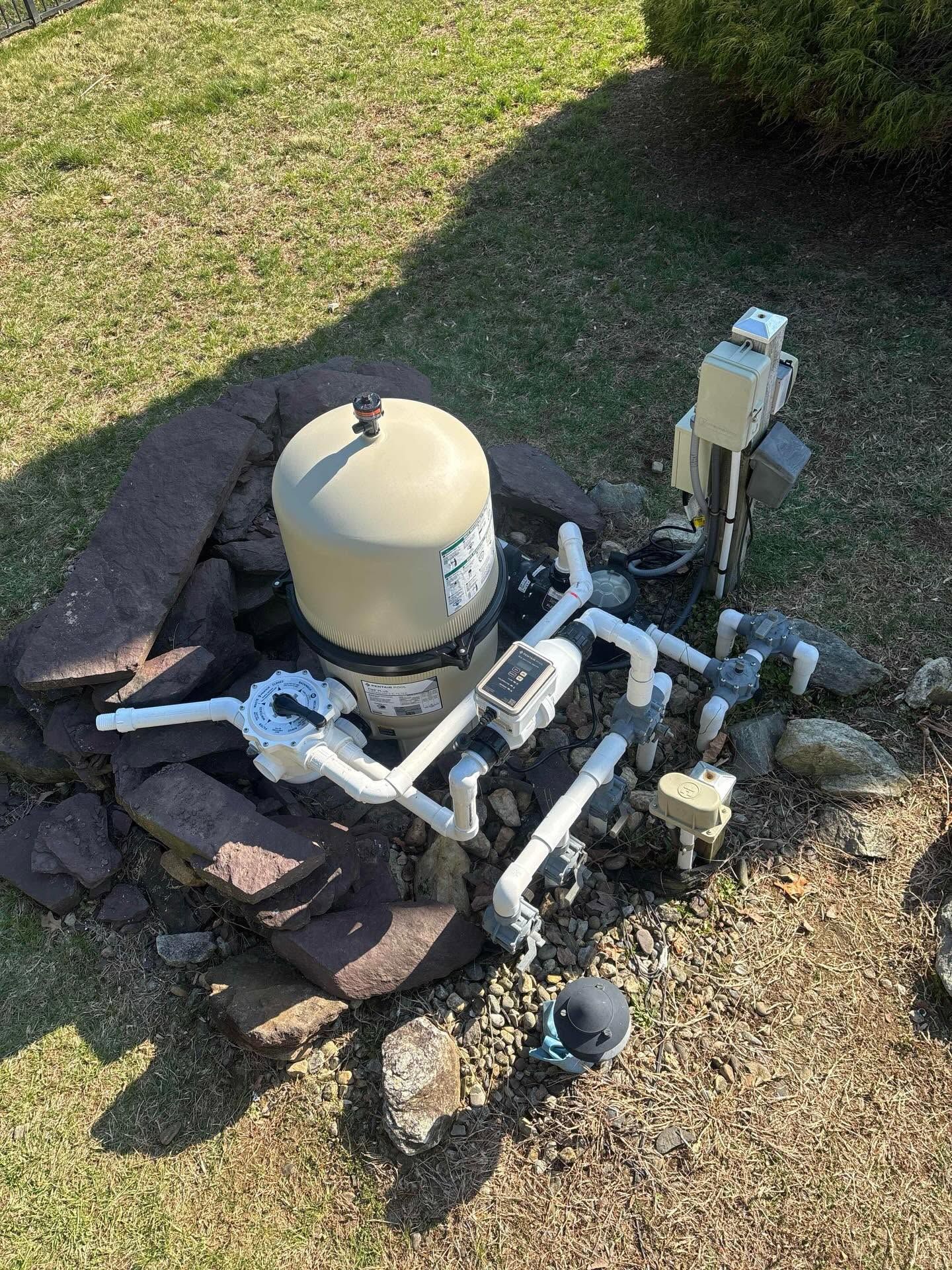 Pool pump and filter equipment with white pipes on a patch of grass next to a wall shadow