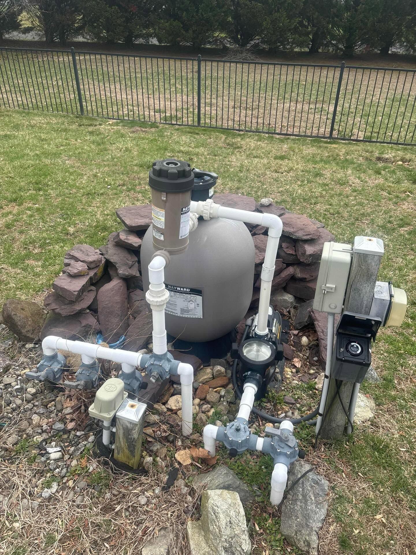 Pool equipment with pipes, pump, and filter beside a rock border in a grassy backyard.