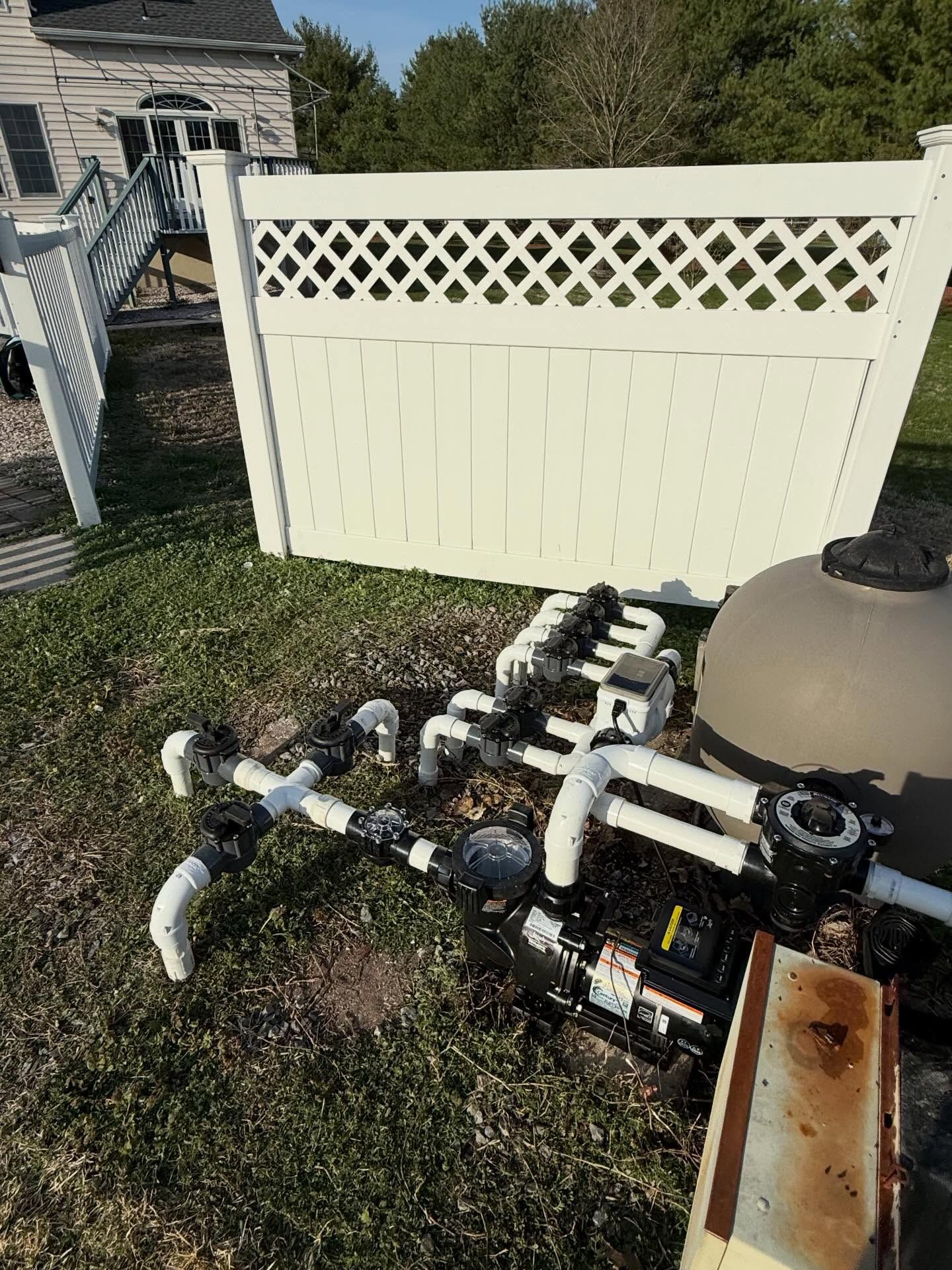 Outdoor pool equipment with white PVC pipes and valves beside a cream privacy fence on grass.