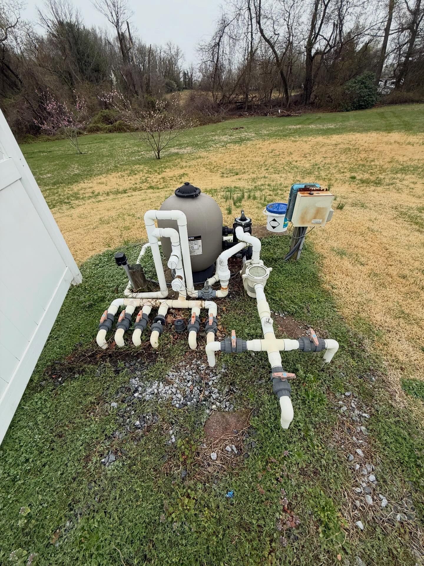 Outdoor pool filtration system with white PVC pipes, a gray tank, and control box on grass near a building.