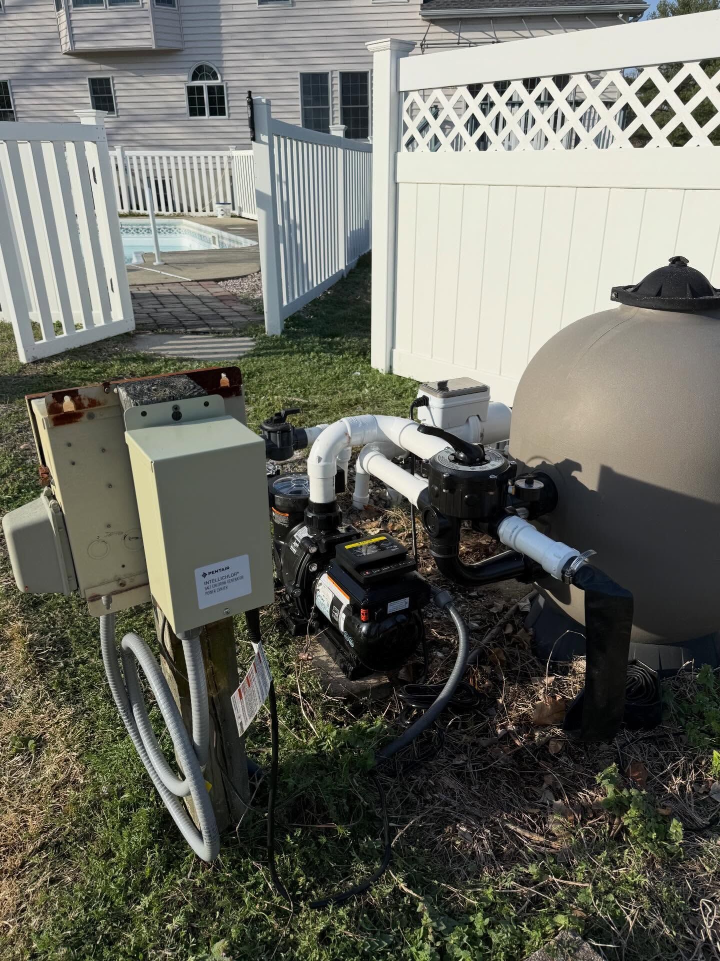Pool pump and filter equipment beside an above-ground pool in a backyard, near a white fence.