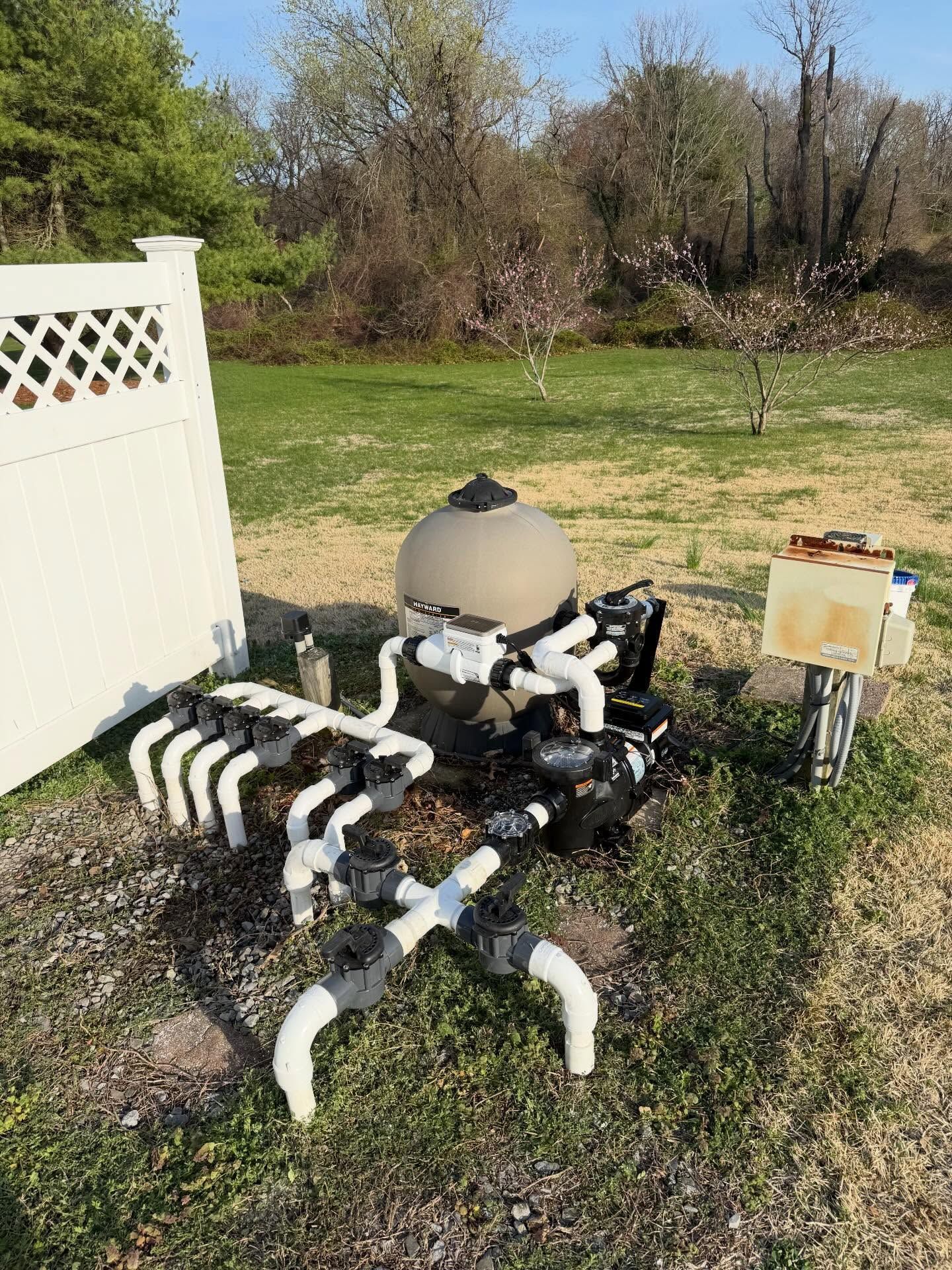 Backyard pool plumbing equipment with white pipes and a beige filter tank on grass.