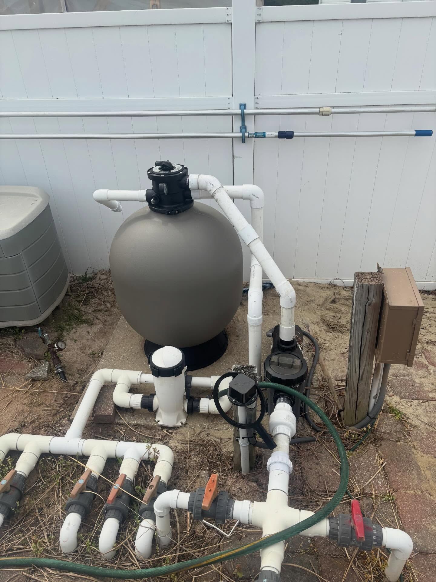 Pool equipment with gray filter tank, pumps, and white PVC pipes beside a fence.