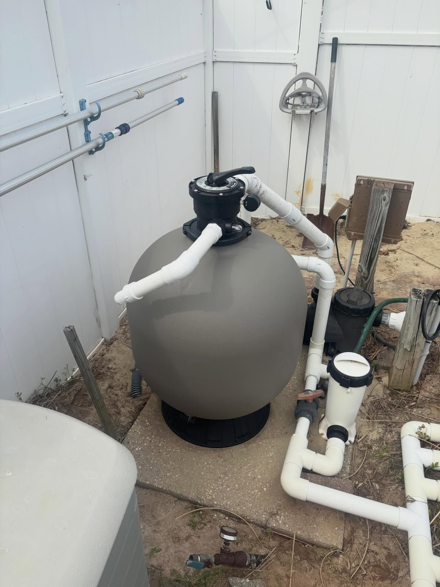 Gray pool filter tank with white pipes and black valve beside an outdoor utility wall