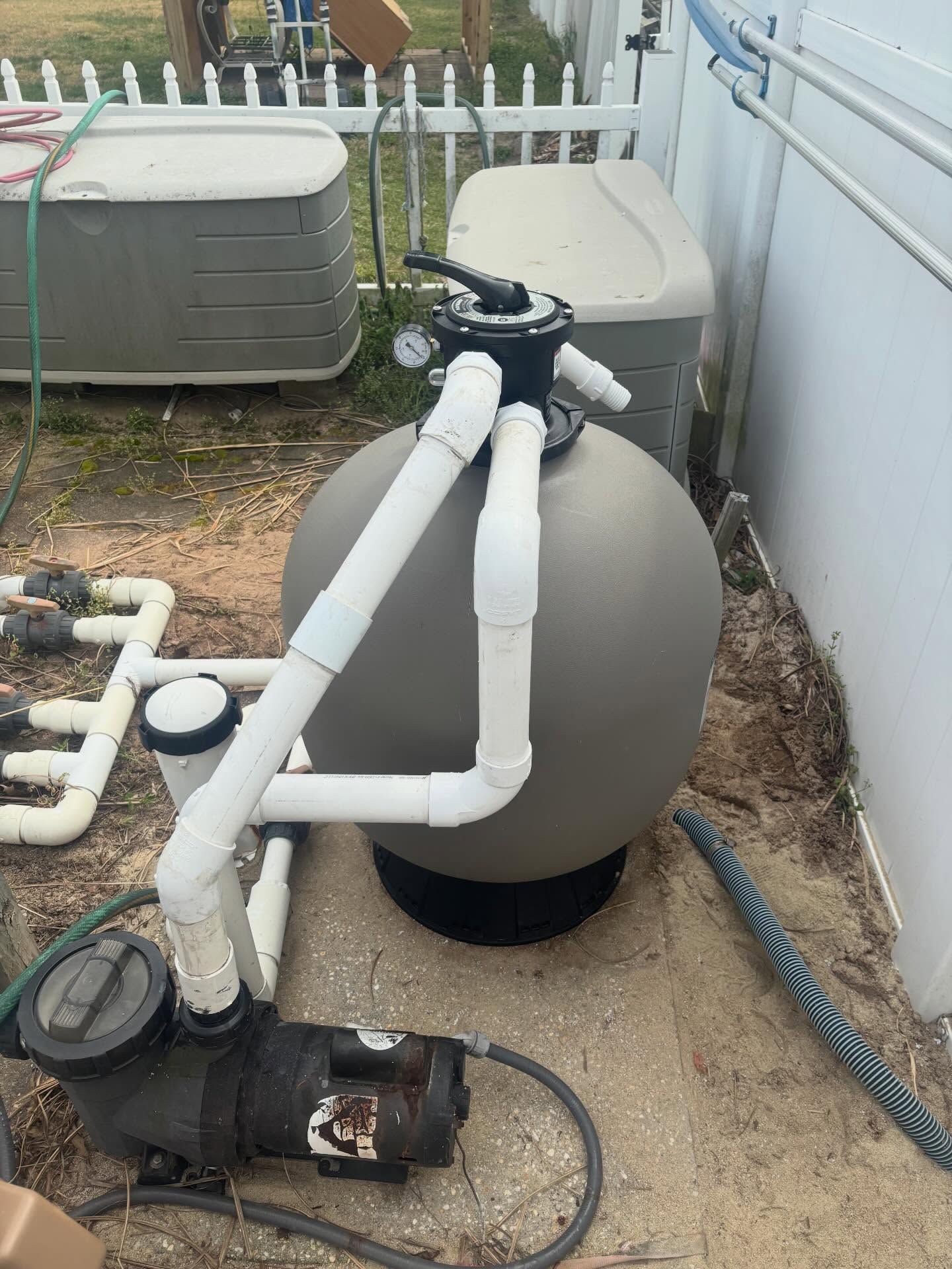Pool equipment with a large gray filter tank, pump, and white pipes beside a house
