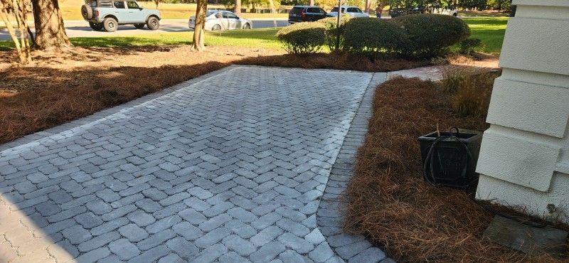 A brick walkway leading to a house with a car parked in the background.