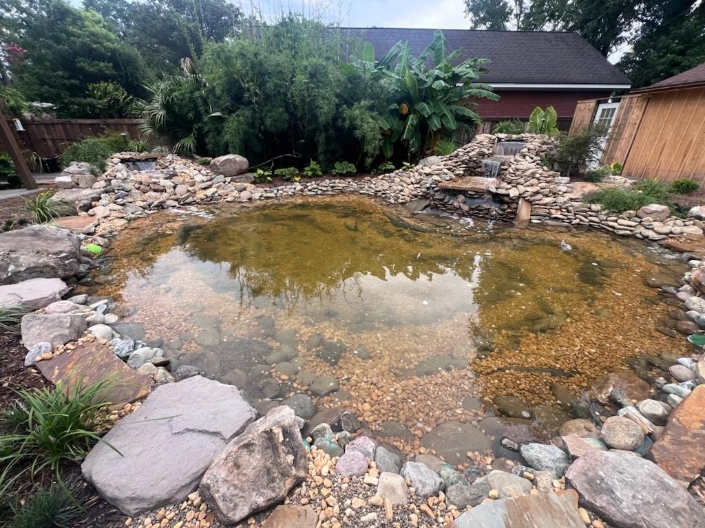 A pond surrounded by rocks and trees in a backyard with a house in the background.