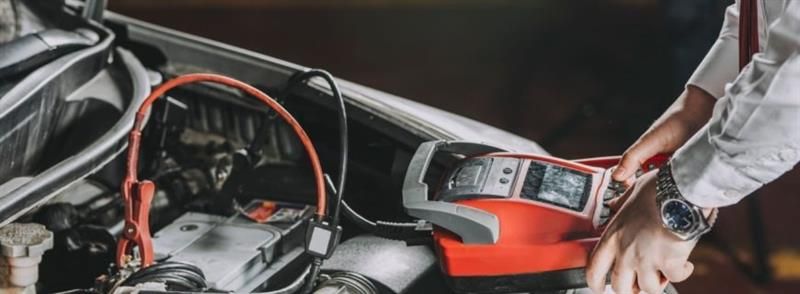 A person uses a red portable battery jump starter to boost a vehicle's engine.