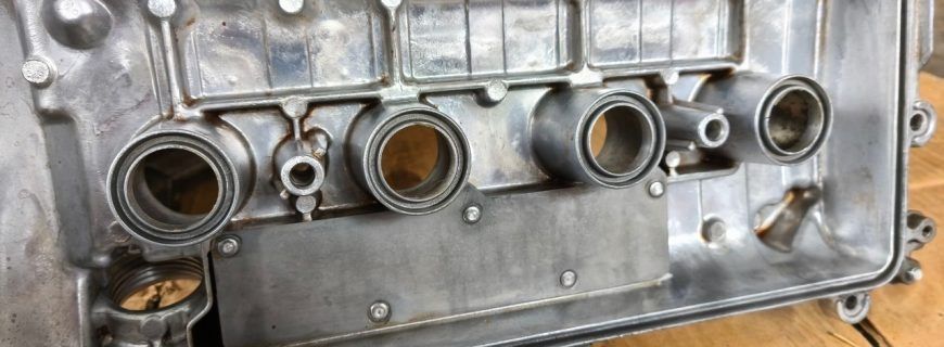 The underside of a metal car engine valve cover showing four cylindrical spark plug tube seals.