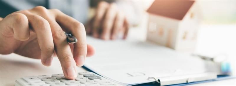 Hands calculating figures on a calculator next to documents and a small house model on a desk.