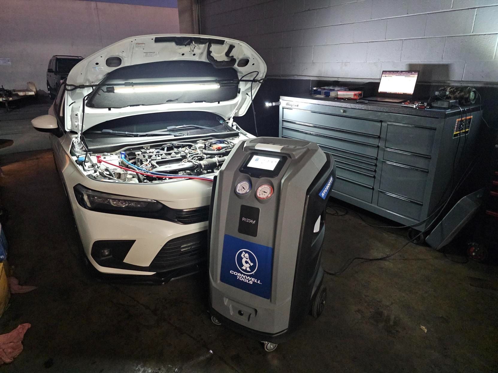 A white car with an open hood is undergoing service in a garage using a nearby A/C refrigerant recovery machine.