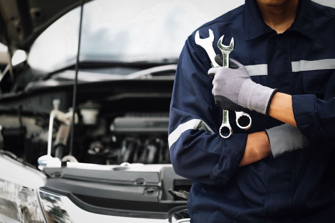 A mechanic in a navy uniform stands with arms crossed, holding two wrenches in front of a car with an open hood.