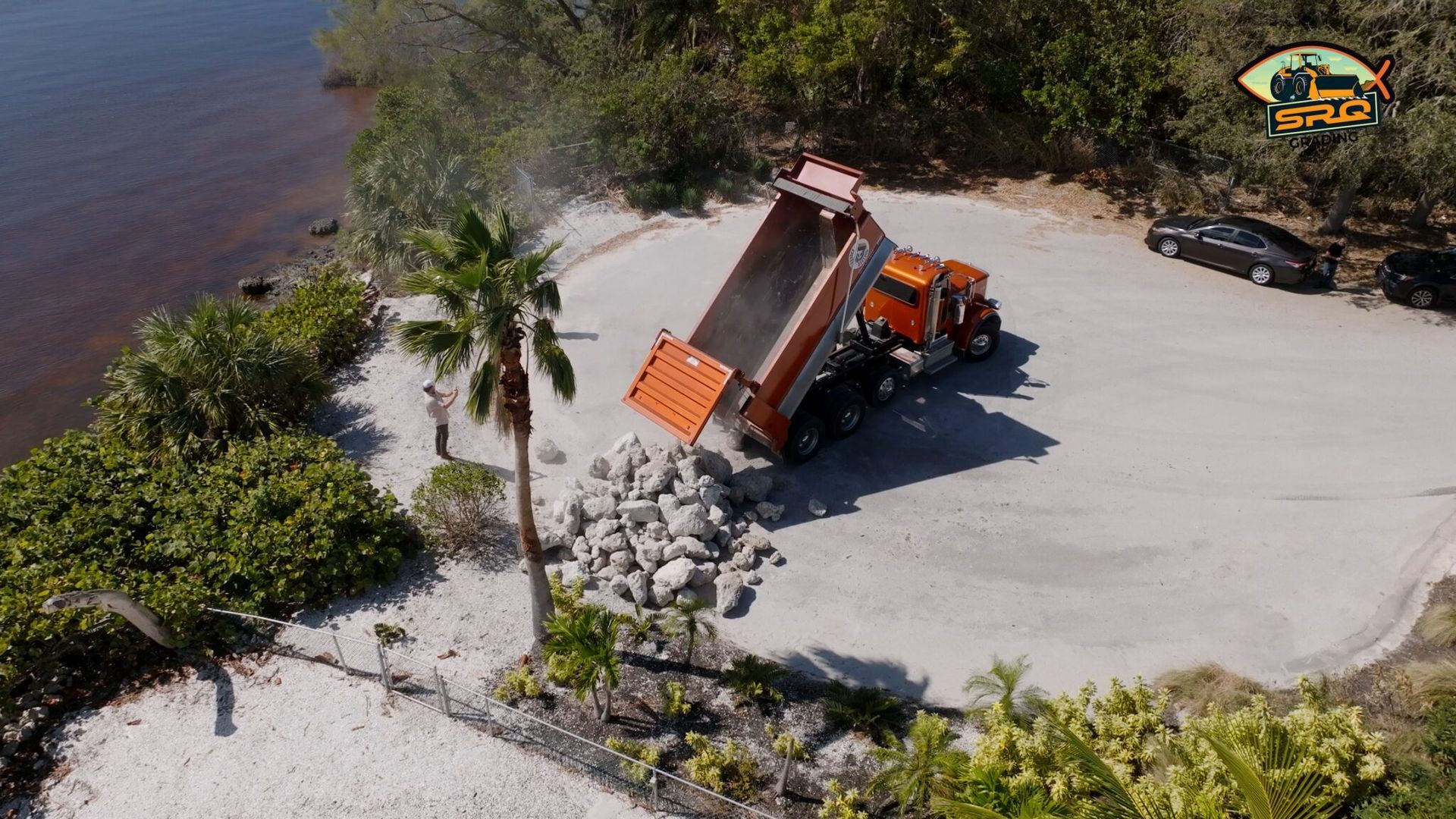 Orange dump truck unloading rocks on a riverside gravel lot, with trees and water nearby