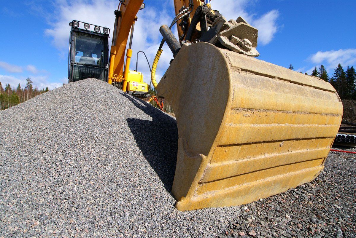 Yellow excavator bucket dumping gravel at a construction site under a blue sky.