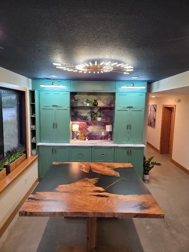 A dining room with a large wooden table, green cabinets, and a colorful mural. Black ceiling with a decorative light fixture.