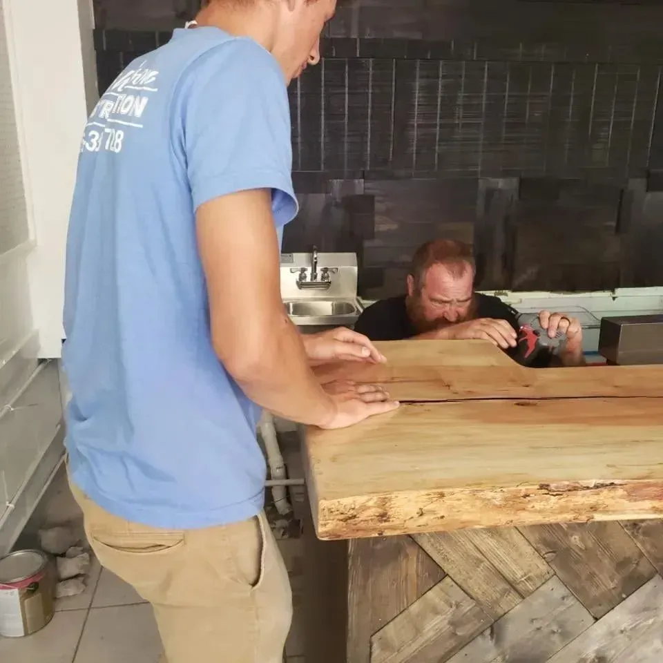 Two men working on a wooden bar counter; one drilling, other pointing, inside a building.