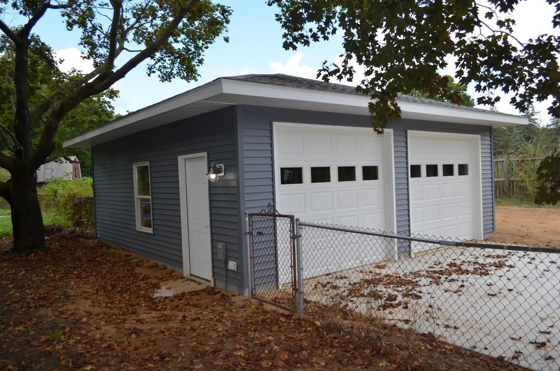 Two-car garage with white doors and gray siding, small white door, chain-link fence, and fall leaves.