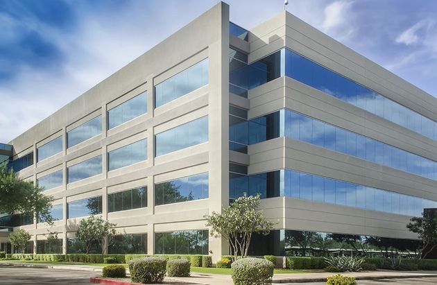 Modern office building with glass windows, beige panels, and surrounding trees under a blue sky.