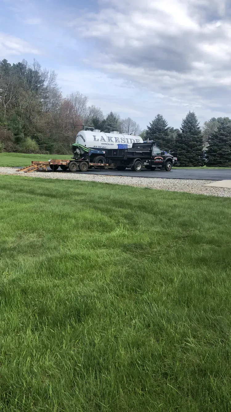 Septic truck parked on driveway; grass in the foreground, trees in the background, overcast sky.