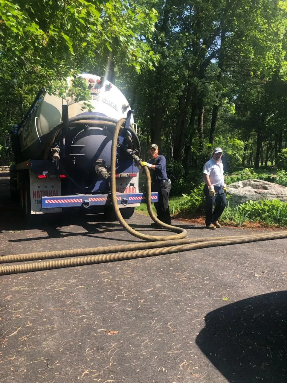 Septic truck with hoses connected, two workers. Trees in the background, asphalt road.