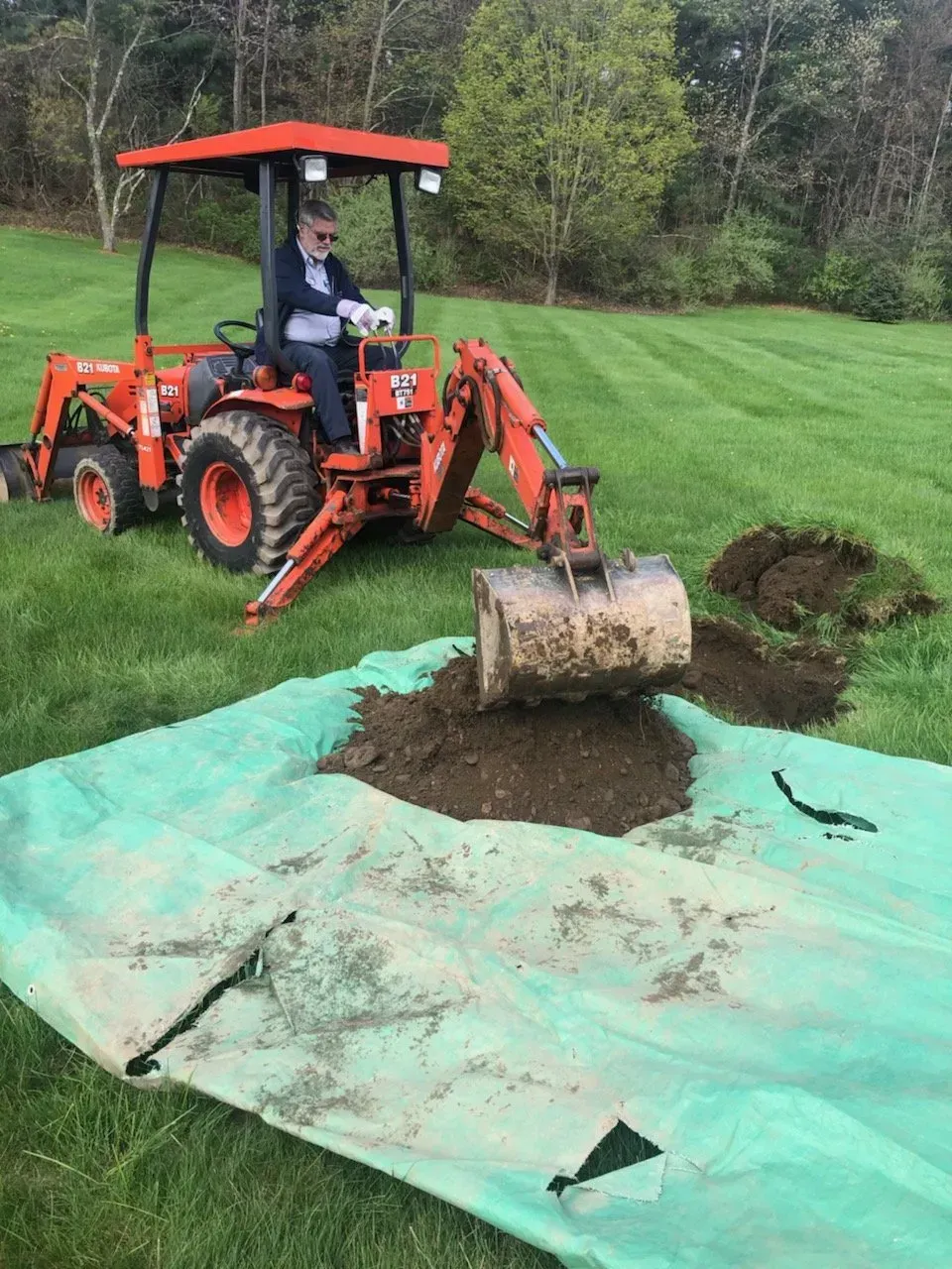 Tractor with backhoe scoops soil onto a green tarp on a grassy lawn. Driver operates the machine.