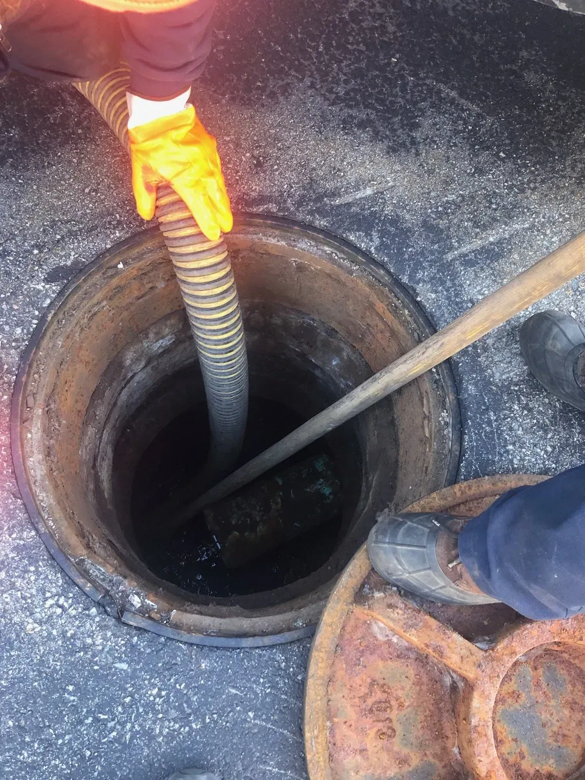 A worker in orange gloves uses a hose to suction liquid from a manhole.