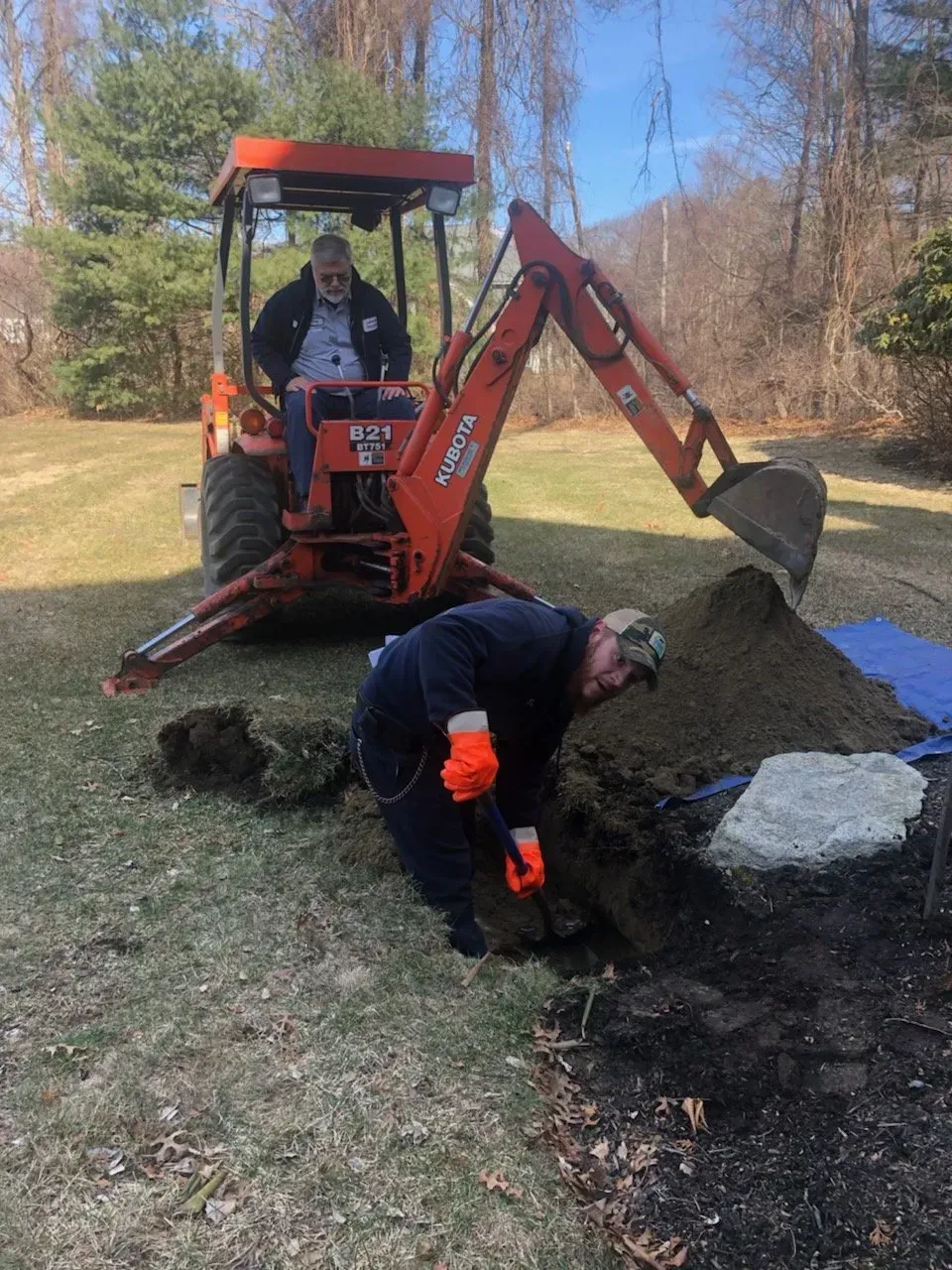 Man operating orange tractor backhoe; another man digging near a soil pile.
