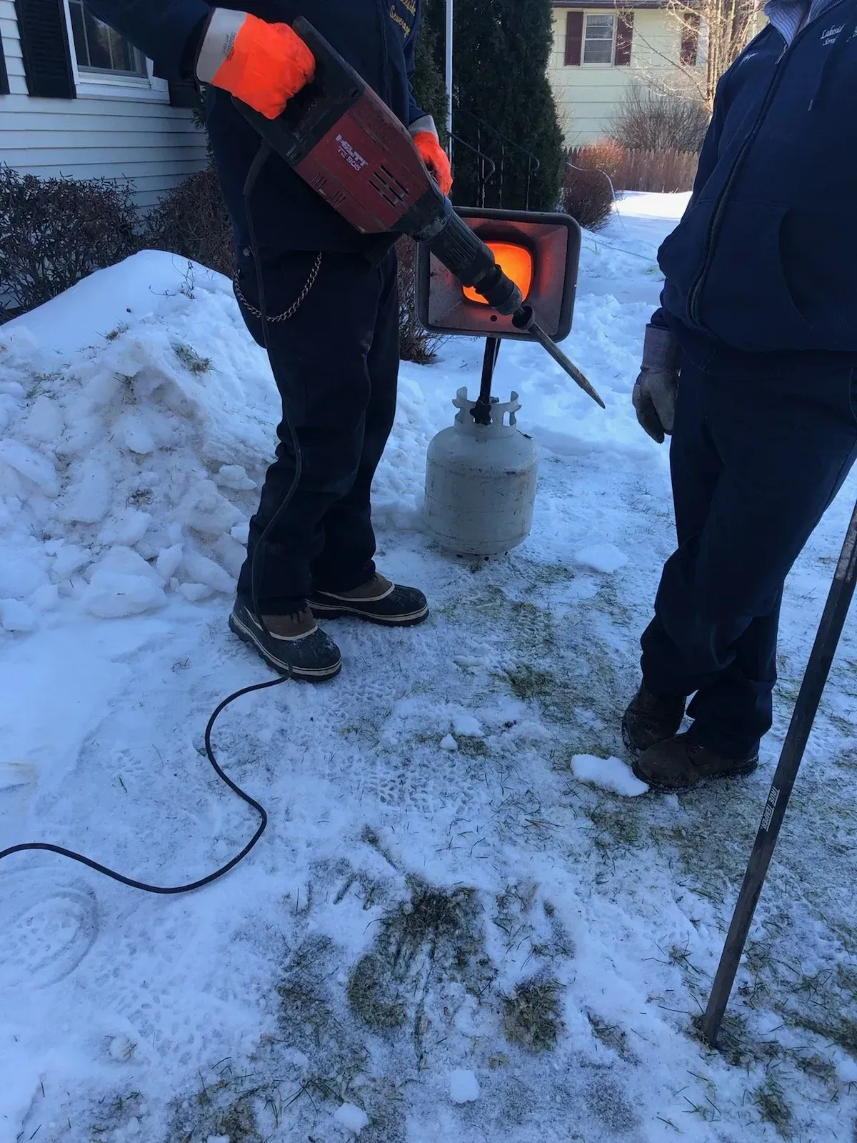 Two people working outdoors with a forge, propane tank, and snow. One holds a jackhammer.