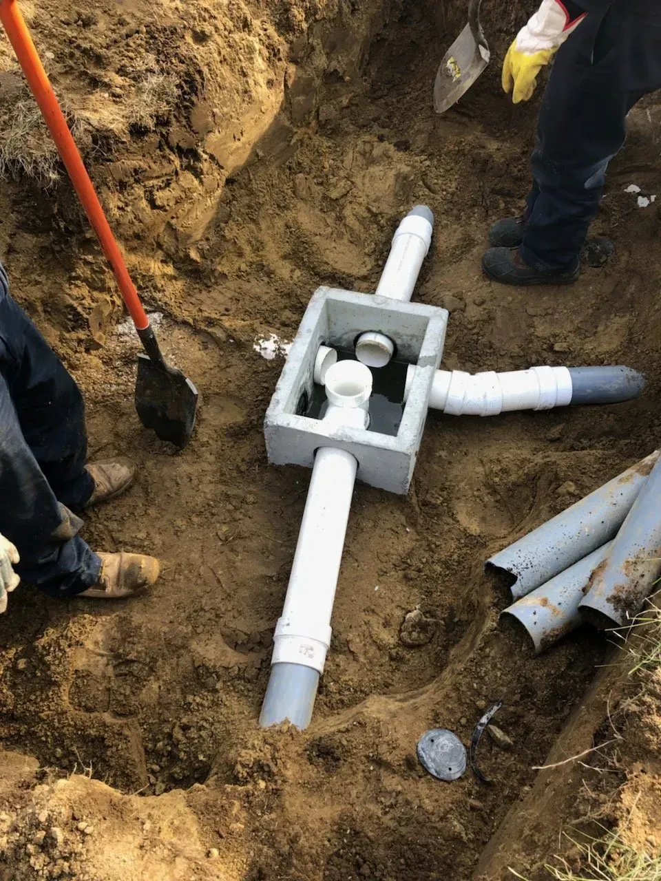 Installation of plumbing fixture in an excavated pit. Workers assemble pipes around a gray concrete junction box.