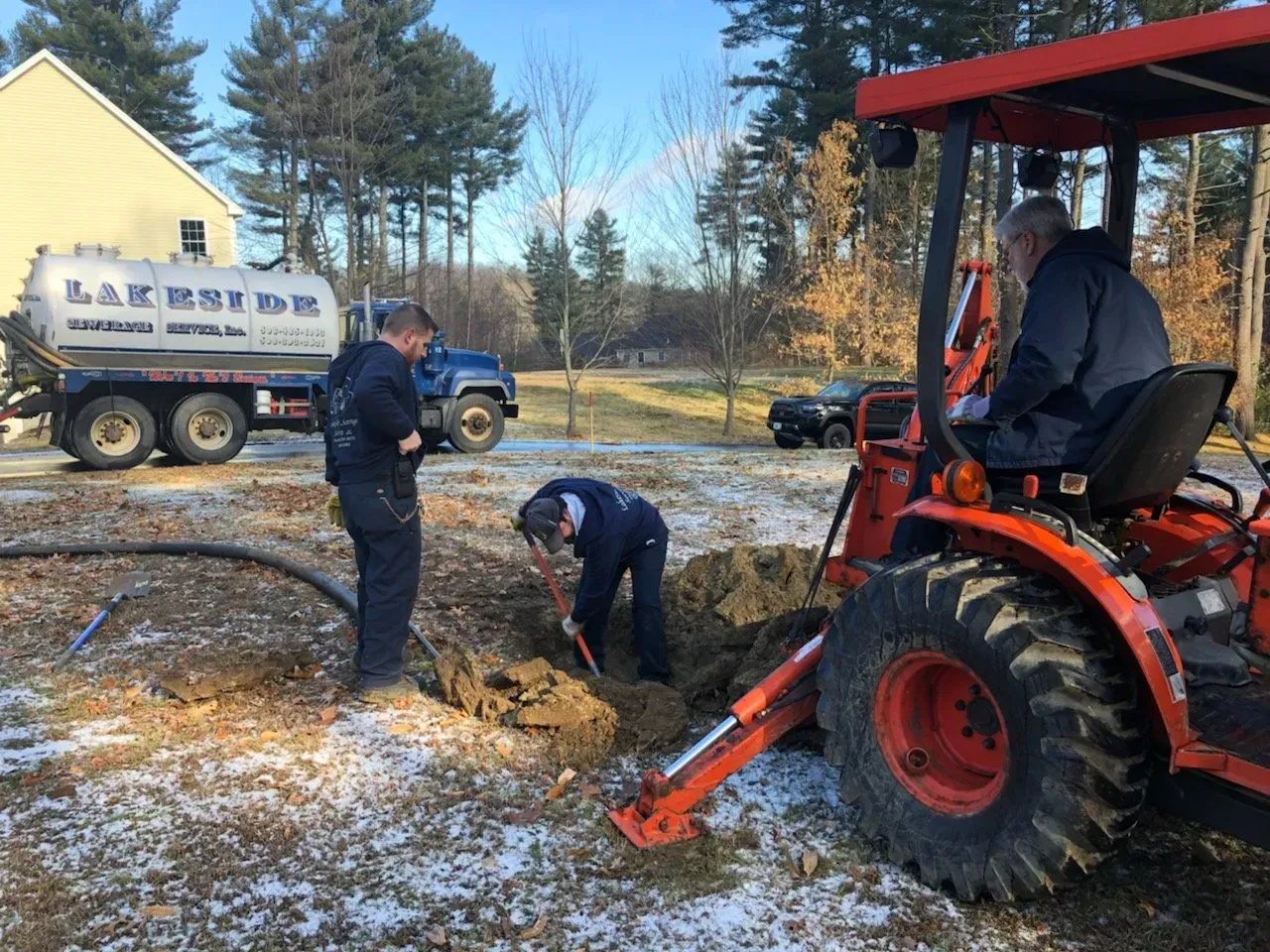 Three men working on a septic system; one drives an orange tractor, two dig in a hole near a septic truck.