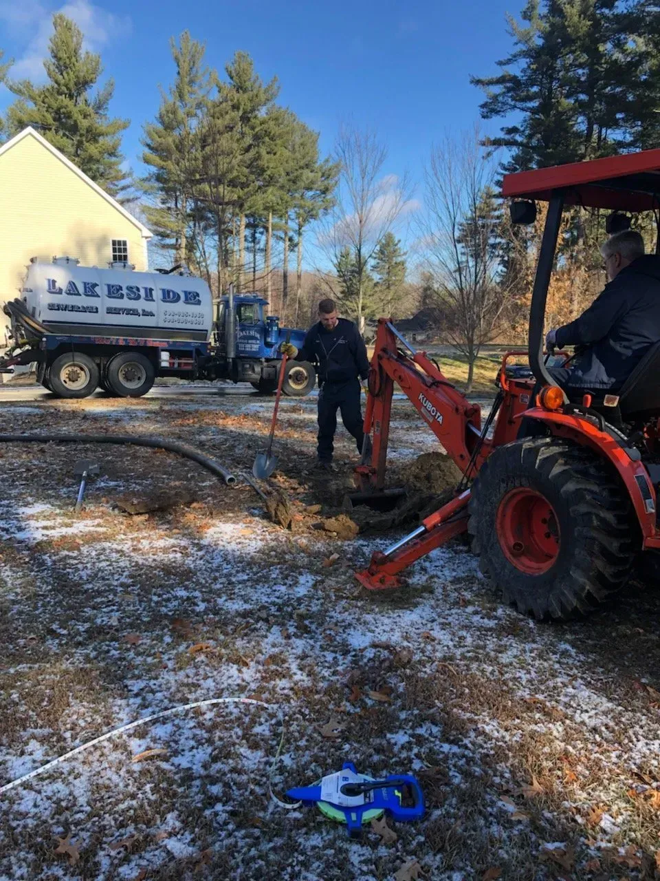 Workers using a tractor and shovel to access a septic tank in a yard, with a service truck nearby.