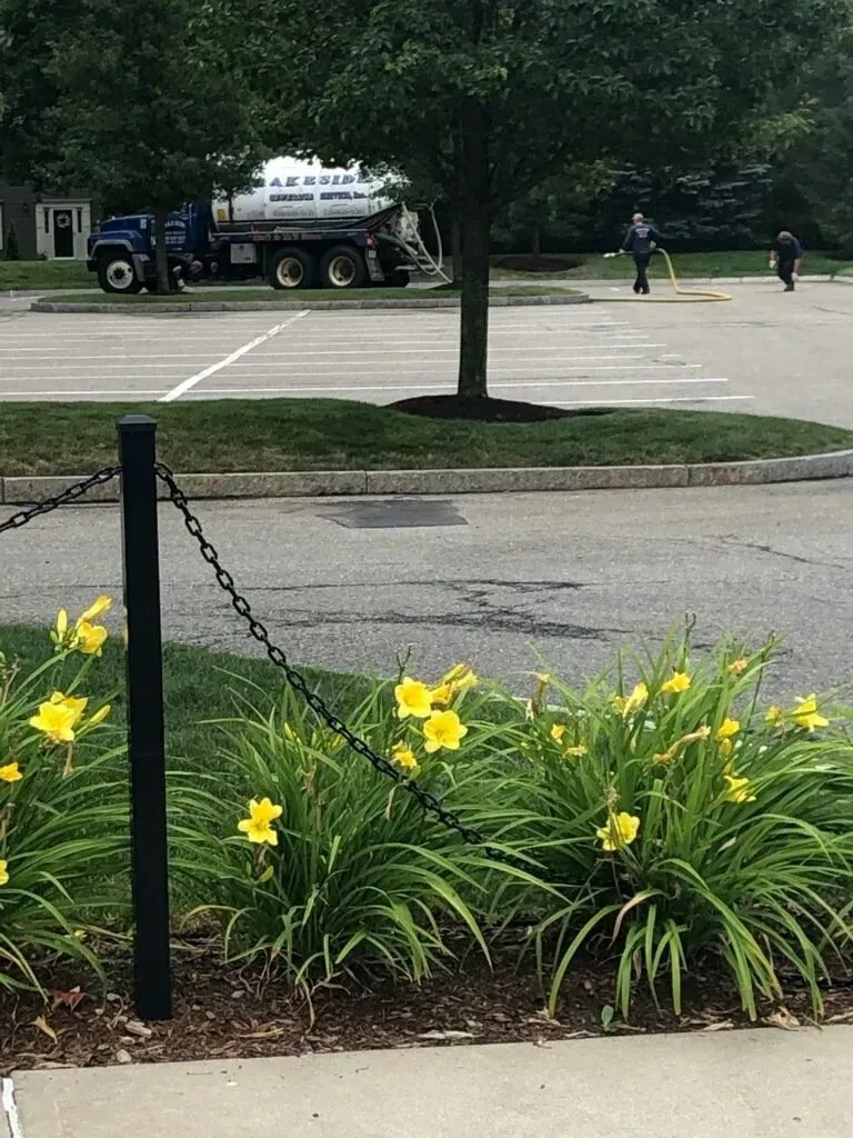 Yellow flowers and a chain fence in the foreground. In the background, a fuel truck and workers in a parking lot.
