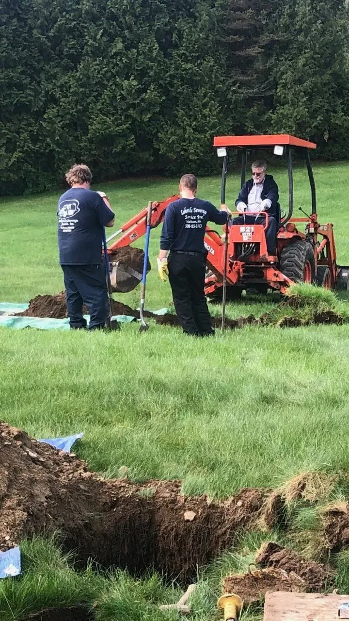 Workers using a backhoe to dig a grave in a grassy field. Two others stand nearby.