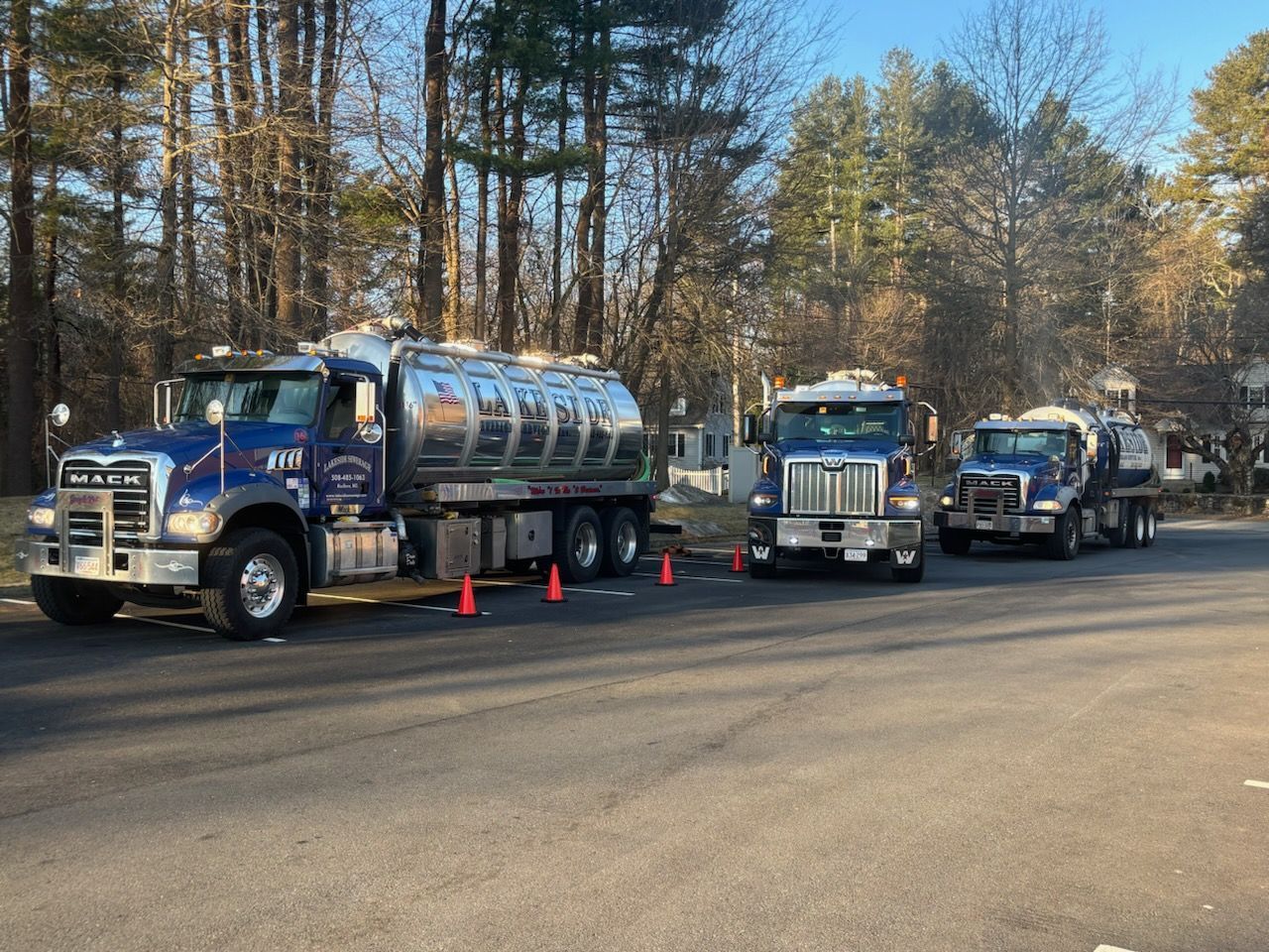 Three tanker trucks parked on a street; trees in background; orange safety cones.