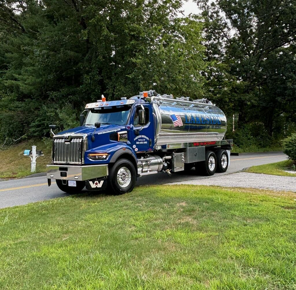 Blue tanker truck on a road, grass in foreground, trees in background, cloudy sky.