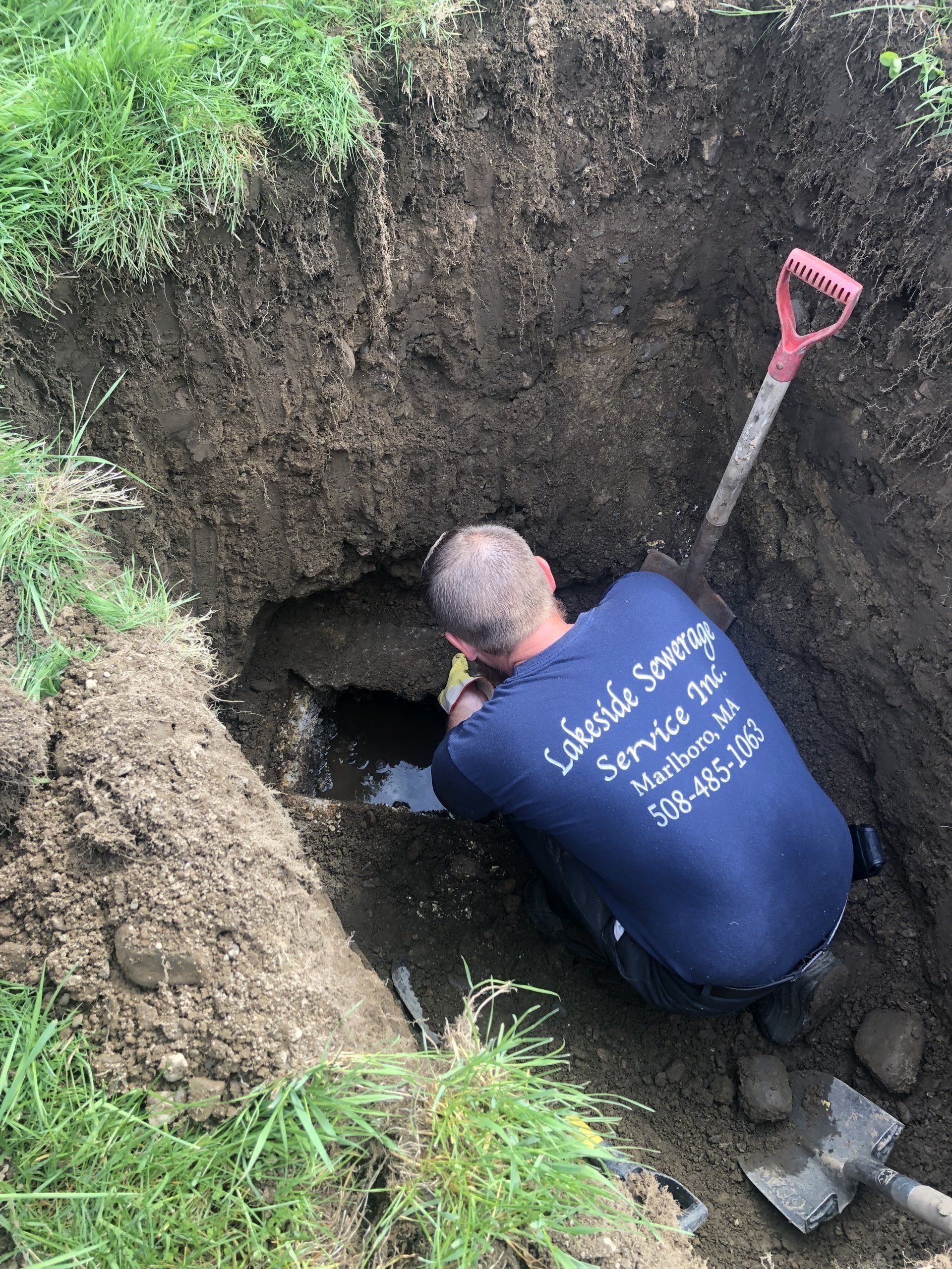 Man in blue shirt, kneeling in a muddy pit, inspecting a pipe. Spade leans against the side.