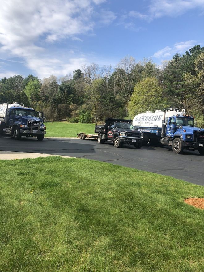 Several trucks parked on asphalt driveway in a grassy yard. Trees in background, blue sky.