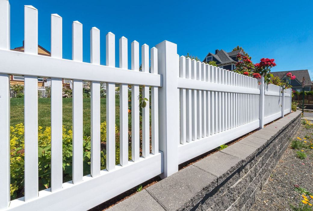 A white picket fence is surrounded by flowers on a sunny day.