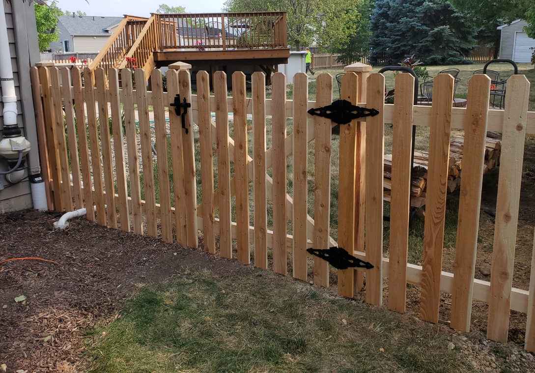 A wooden picket fence with a gate and a deck in the background.