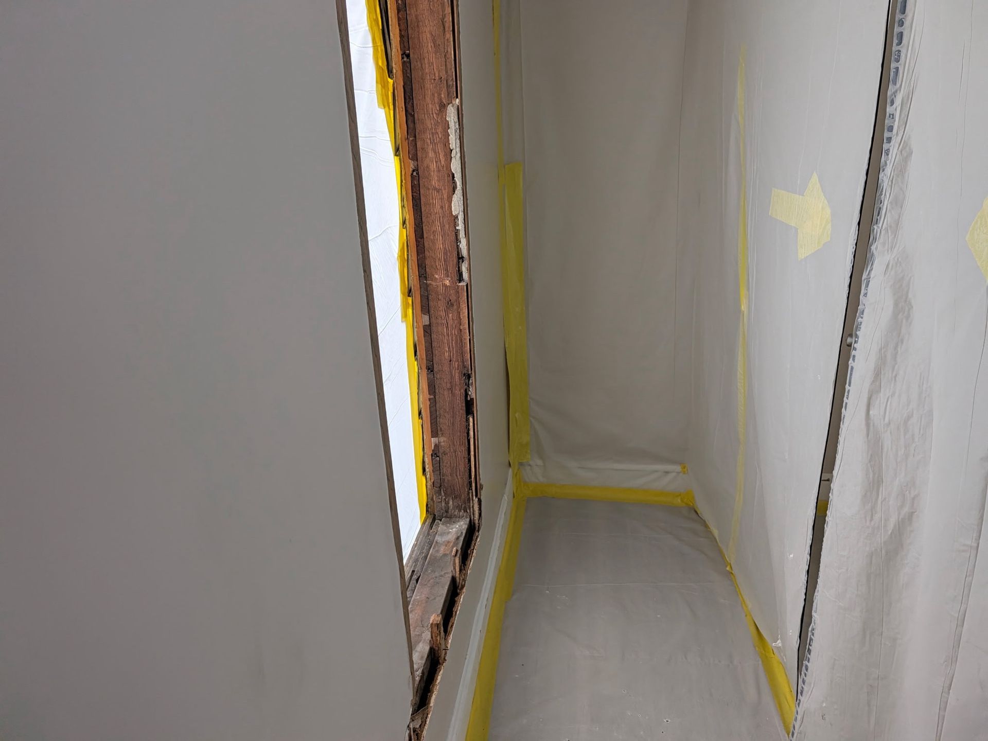 Narrow hallway under construction with plastic sheeting, yellow tape, and a window opening on the left.