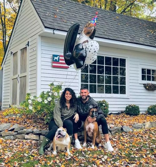 Couple with three dogs sitting outside a white house, one wearing a party hat and float ring