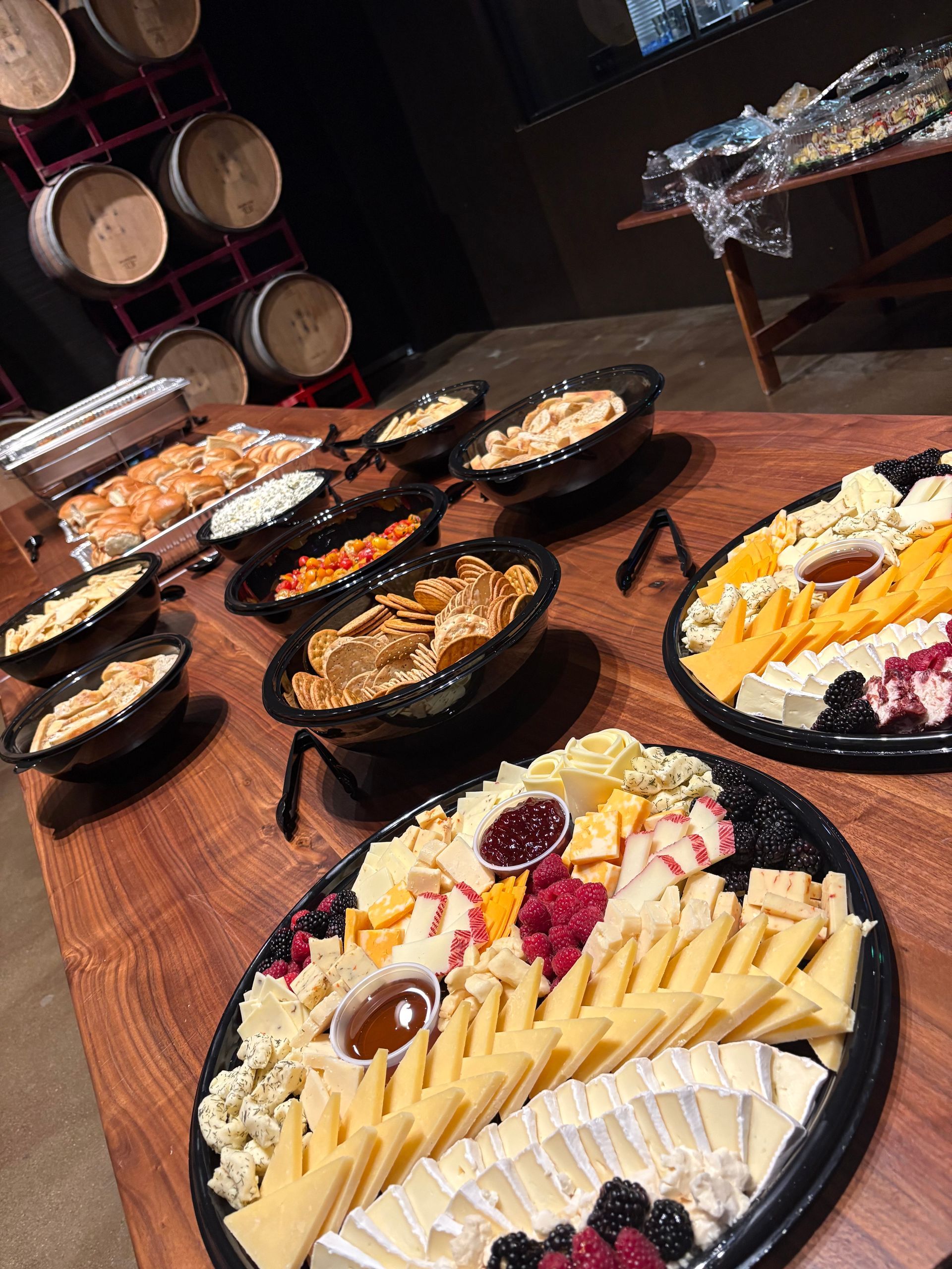 Buffet spread on a wooden table, featuring cheese platters, dips, and warm food. Barrels in background.