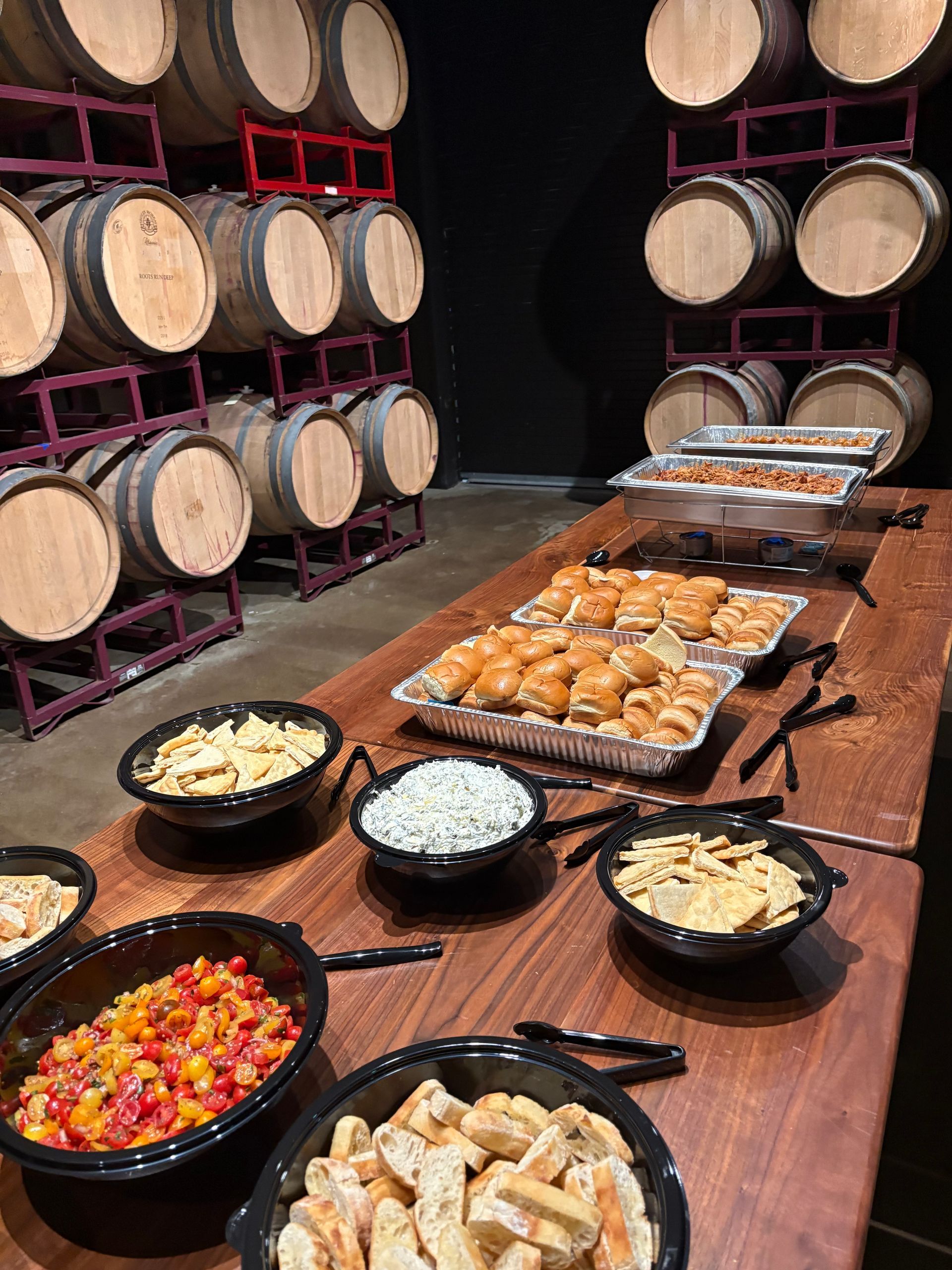 Buffet table with various foods in front of wine barrels in a dimly lit setting.