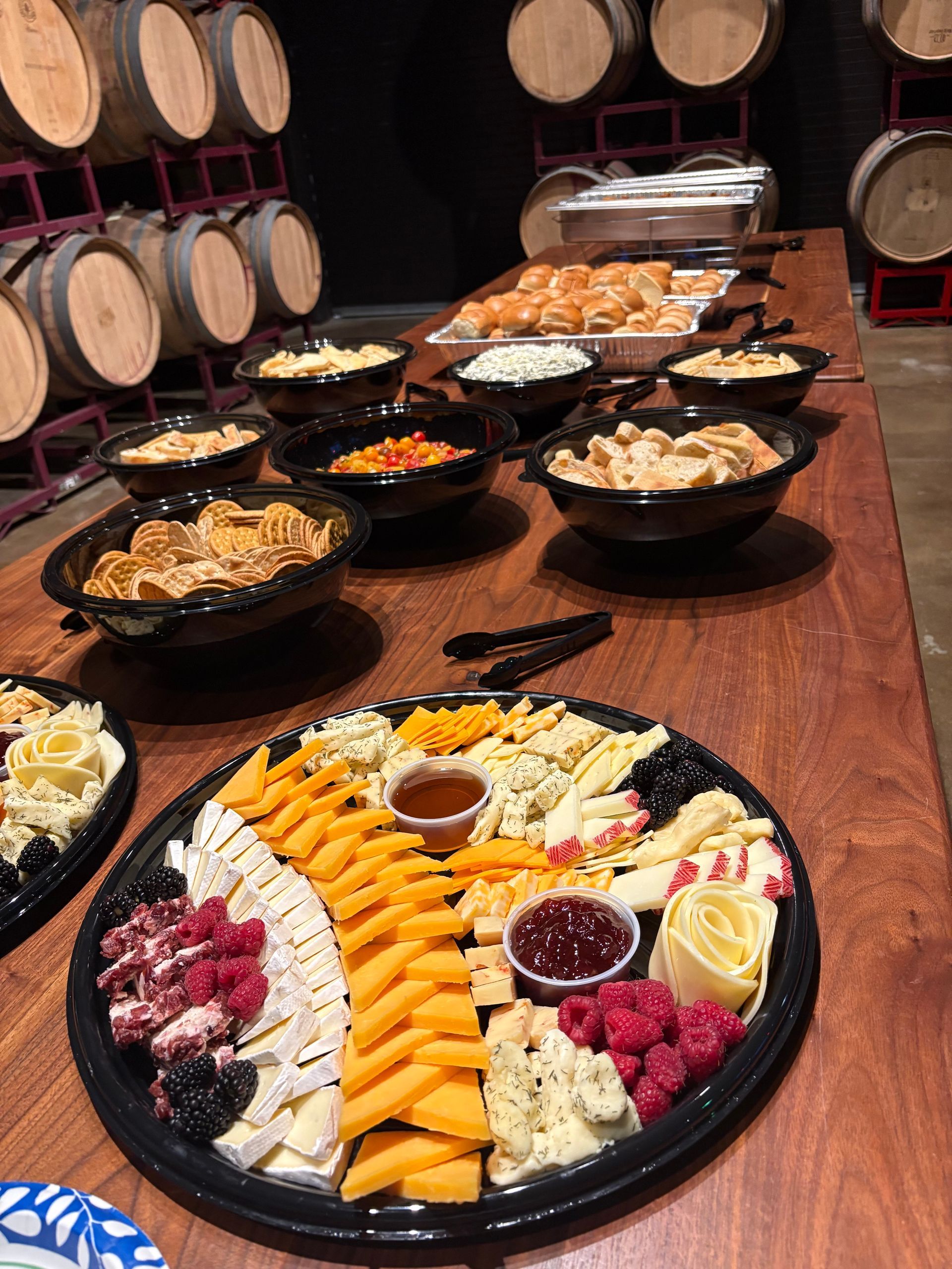 Buffet table with cheese platter, crackers, dips, and small rolls, barrels in background.