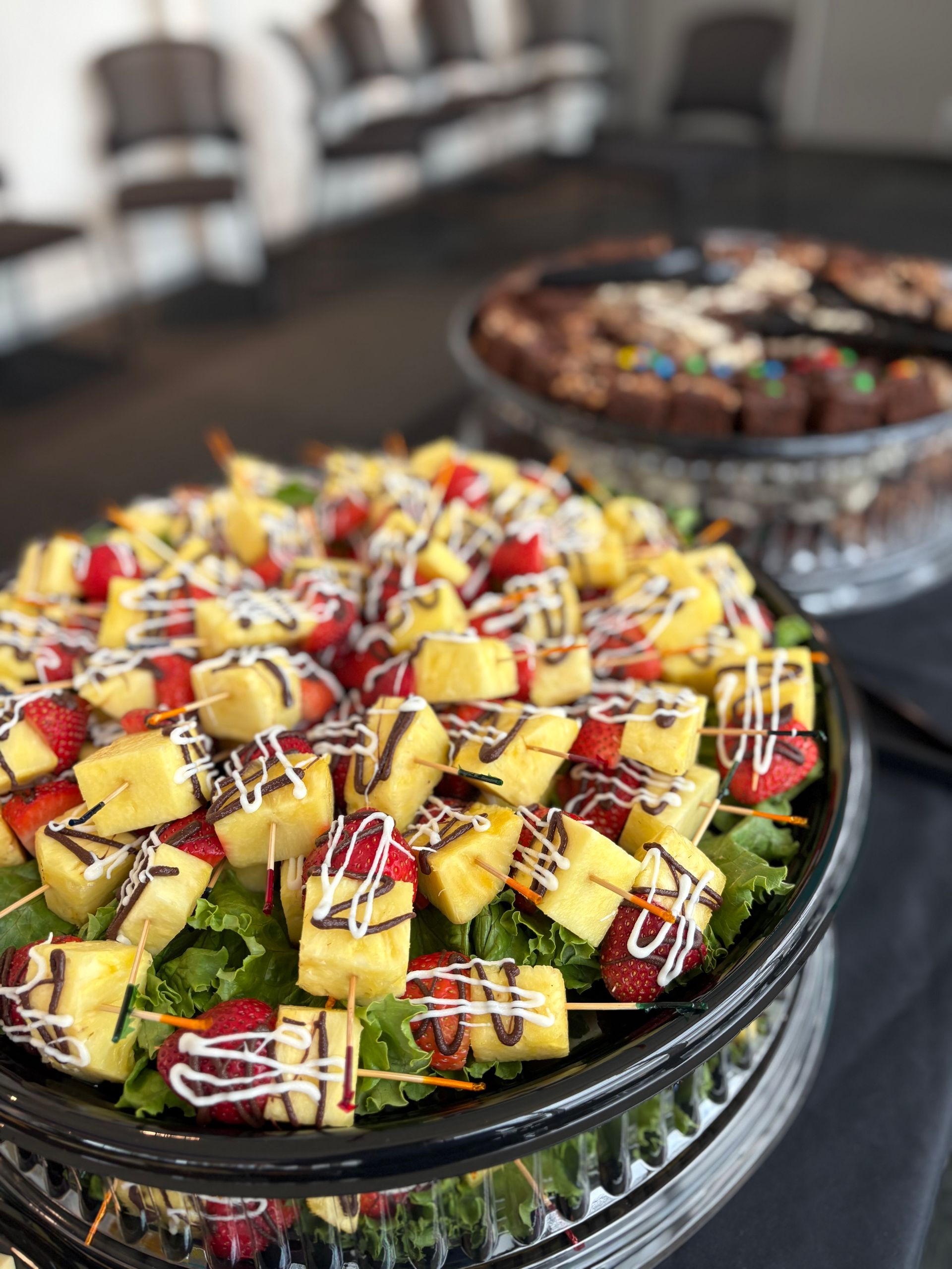Fruit skewers with chocolate drizzle on a platter, and a tray of brownies in the background.