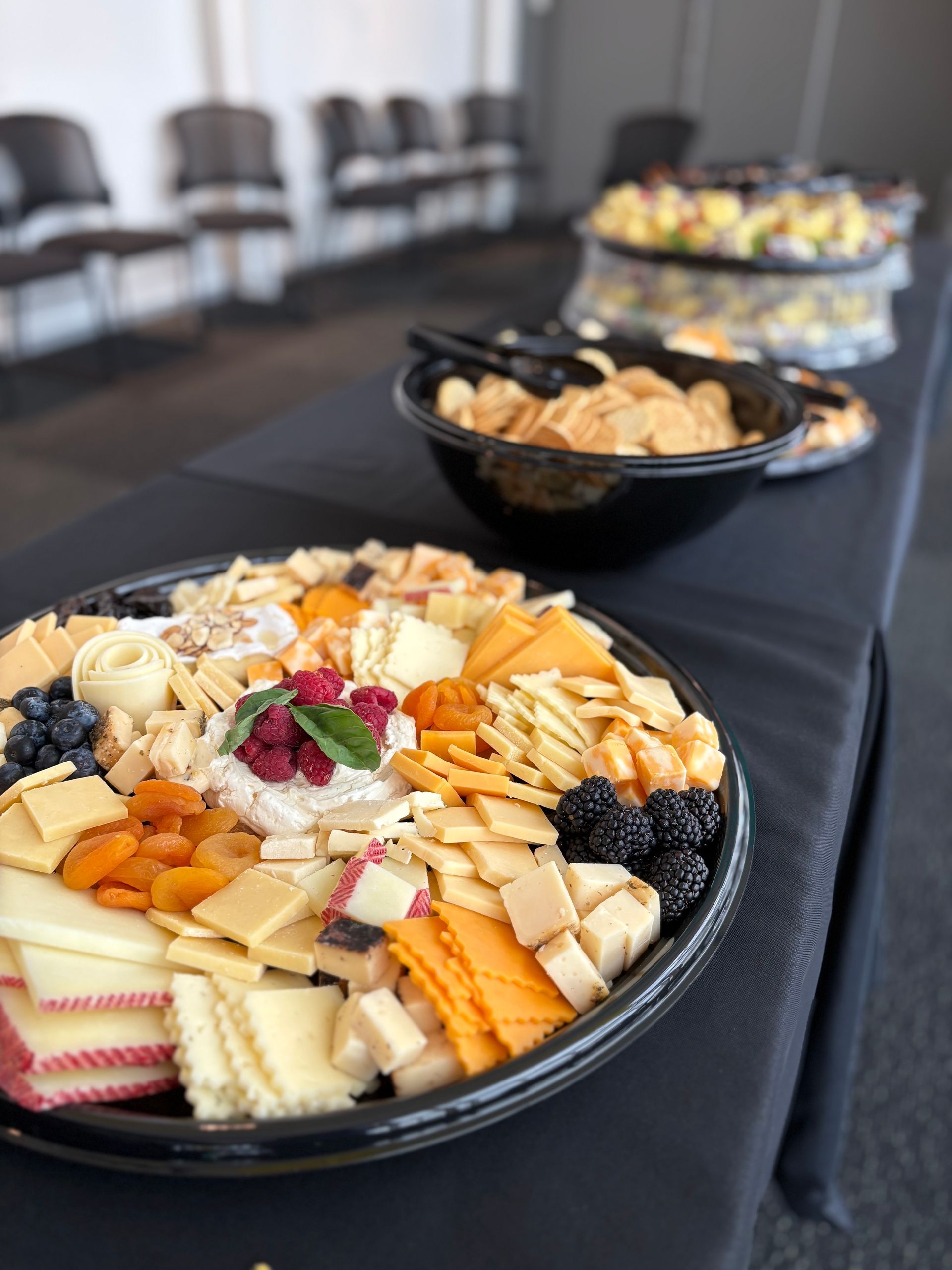 A platter of cheeses, fruits, and crackers on a black table. Other snacks and chairs in background.